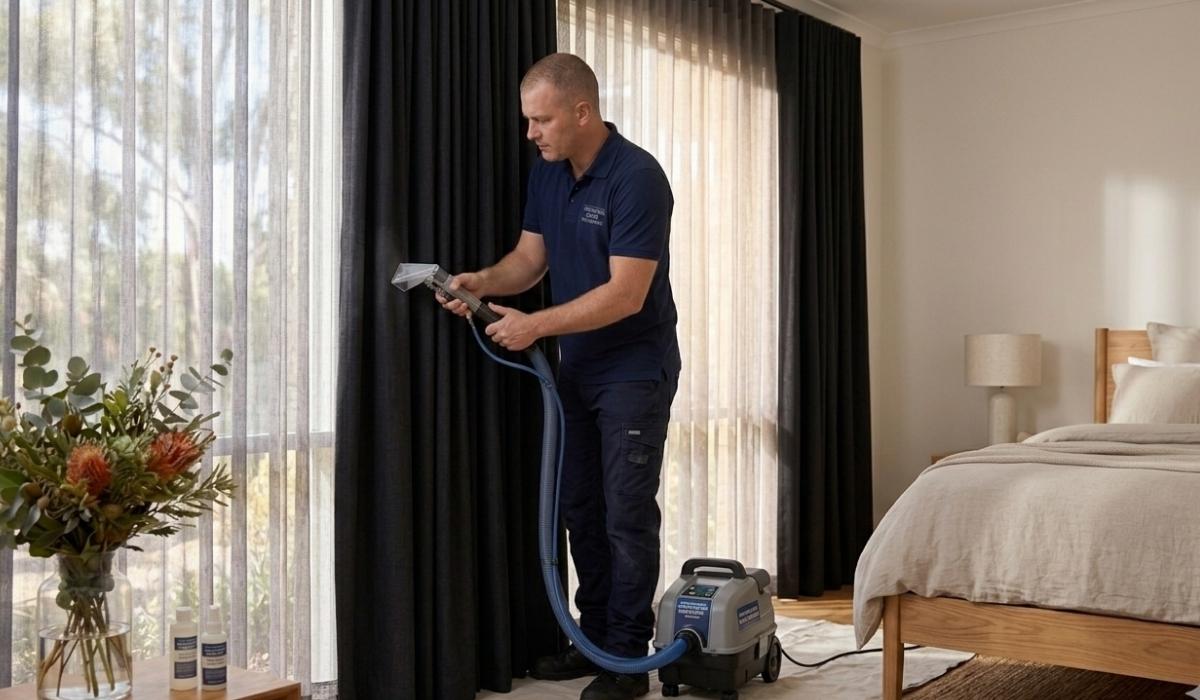 A technician in a navy uniform uses a specialized steam attachment to clean dark blackout curtains in a modern, sunlit bedroom.
